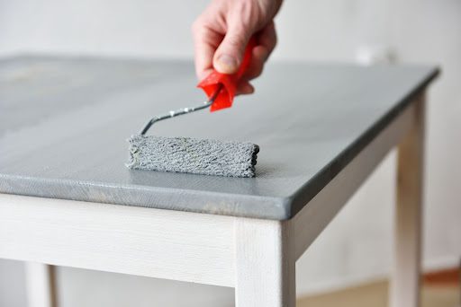 A handheld paint roller is spreading slate gray paint atop a white table.