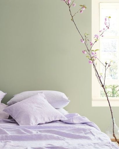 A bedroom showcasing interior color trends. It has a pale green color on the wall and an unmade bed with purple sheets.